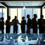 A silhouette of a line of co-workers standing and talking with each other in front of a high-rise window.