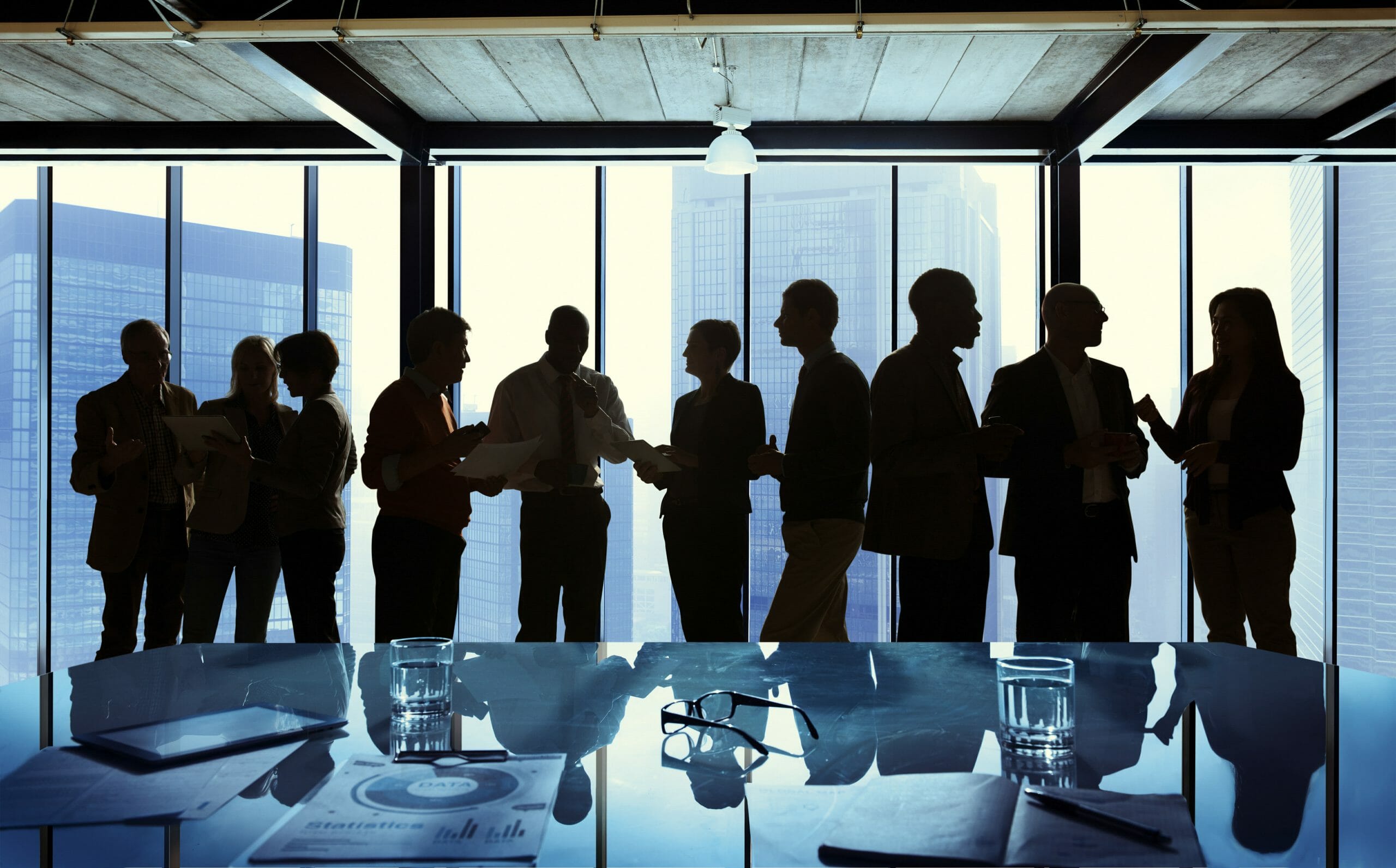 A silhouette of a line of co-workers standing and talking with each other in front of a high-rise window.