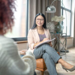 Pleasant psychologist wearing grey trousers smiling to client while speaking with her